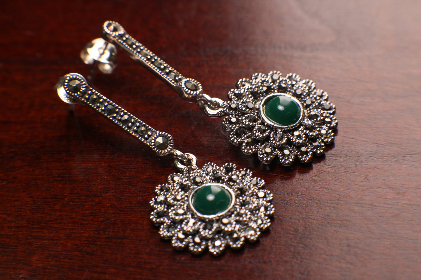 May include: A pair of silver dangle earrings with green gemstones. The earrings feature a delicate floral design with a round green gemstone in the center of each flower. The earrings are made of silver and have a polished finish.