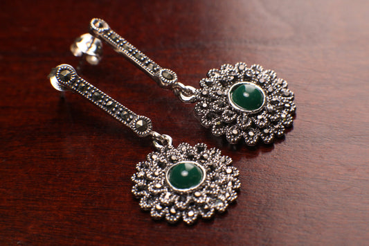 May include: A pair of silver dangle earrings with green gemstones. The earrings feature a delicate floral design with a round green gemstone in the center of each flower. The earrings are made of silver and have a polished finish.