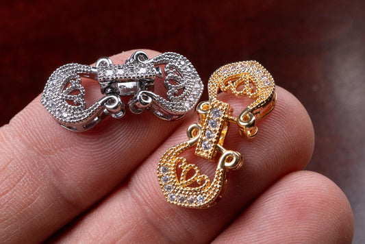 May include: Close-up of two ornate jewelry clasps. One is silver-toned, the other gold-toned. Both clasps are encrusted with small, clear gemstones. The clasps have a decorative, openwork design with curved elements.